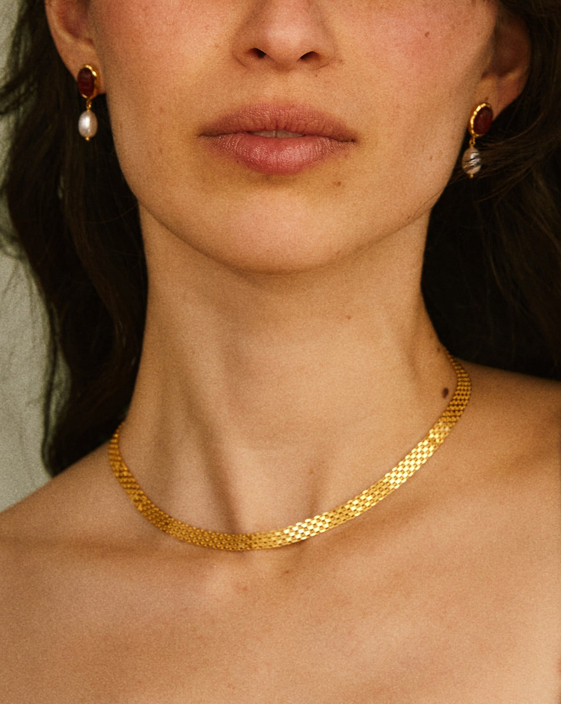 Close-up of a person wearing a gold necklace and earrings with a neutral background