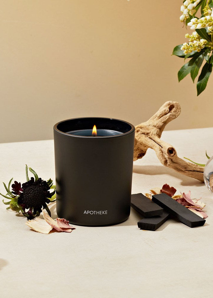 Black candle with a lit wick on a beige surface, surrounded by flowers and driftwood.