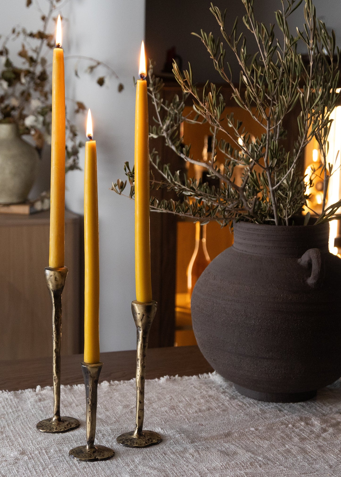Three tall yellow candles in decorative holders on a textured surface with a vase and branches in the background.