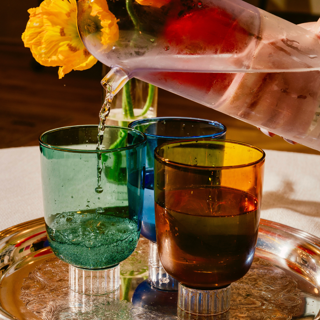 Hand pouring a colorful drink into three differently colored glasses on a reflective surface.