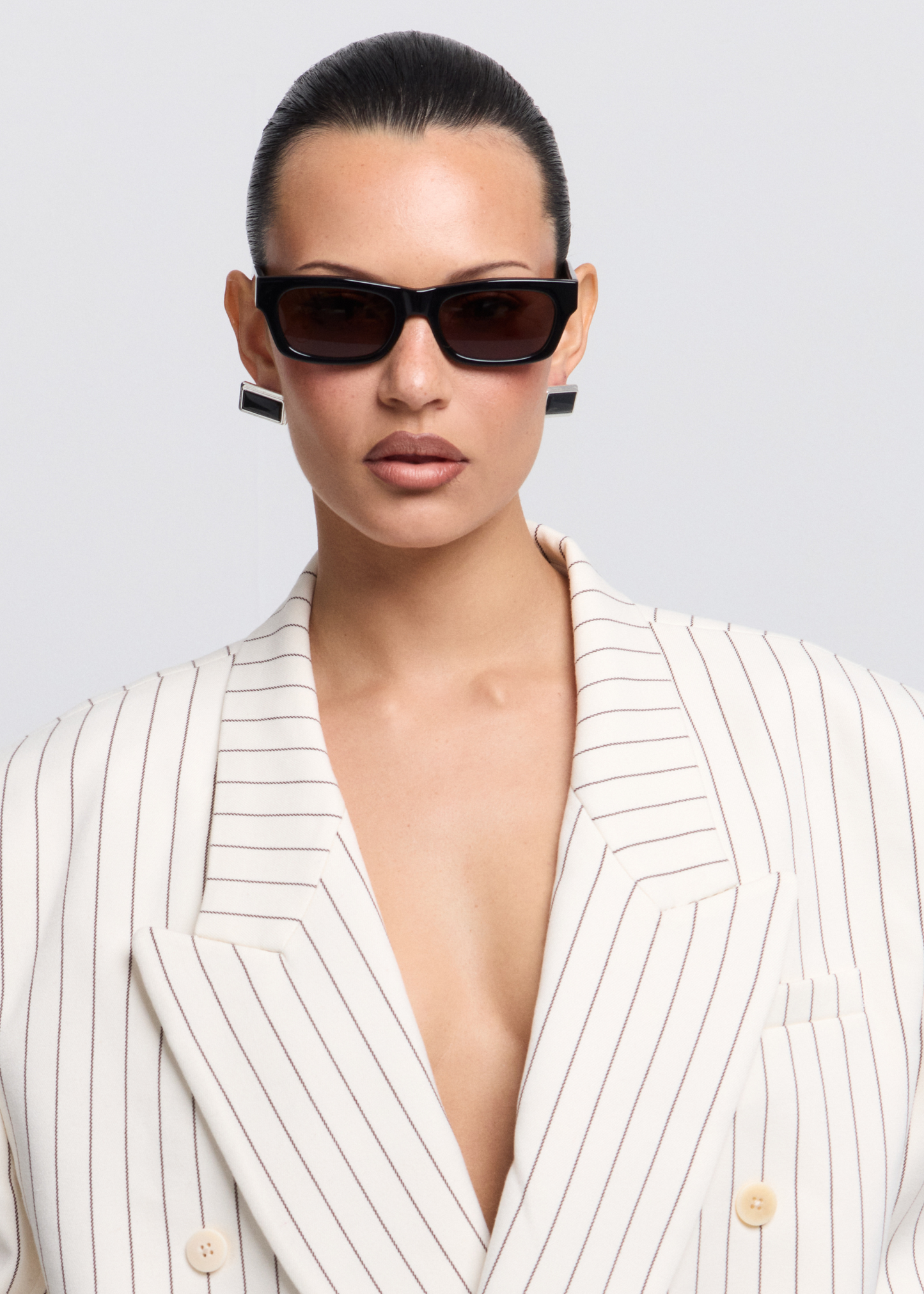Woman wearing black sunglasses, a white pinstripe blazer with a low neckline against a light gray background