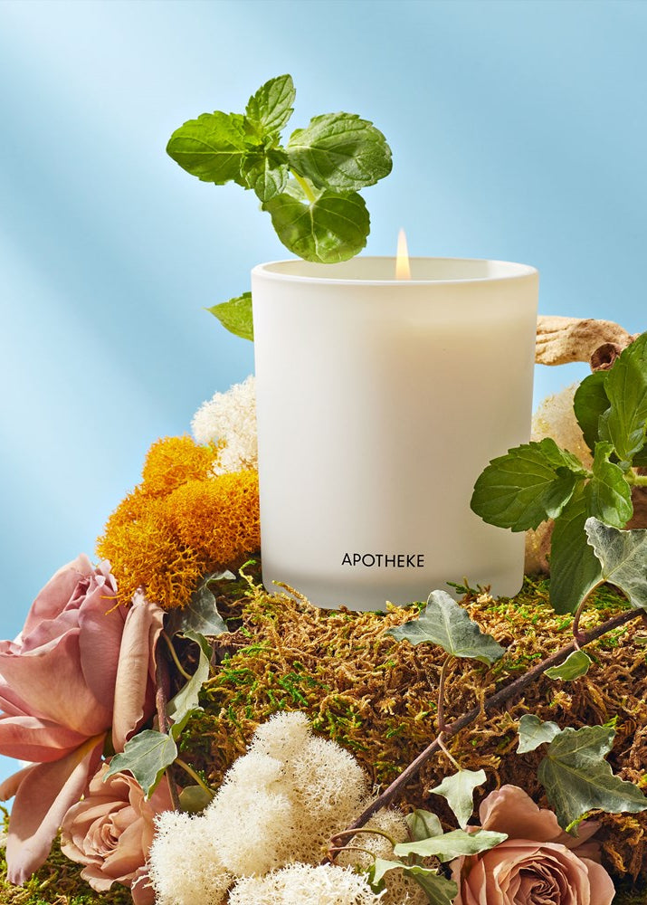 White candle labeled 'Apotheke' surrounded by flowers and greenery on a blue background