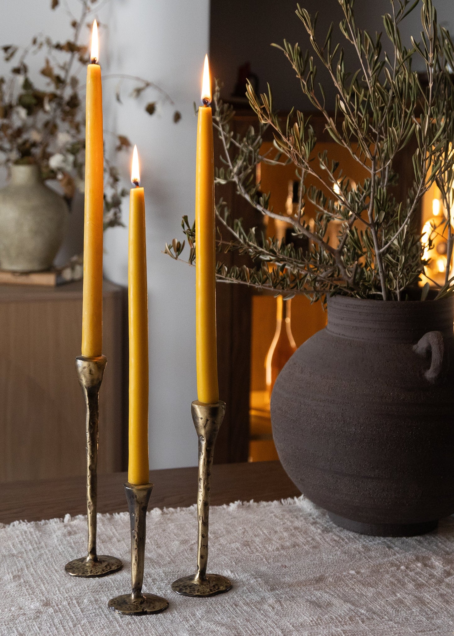 Three tall yellow candles in decorative holders on a textured surface with a vase and branches in the background.