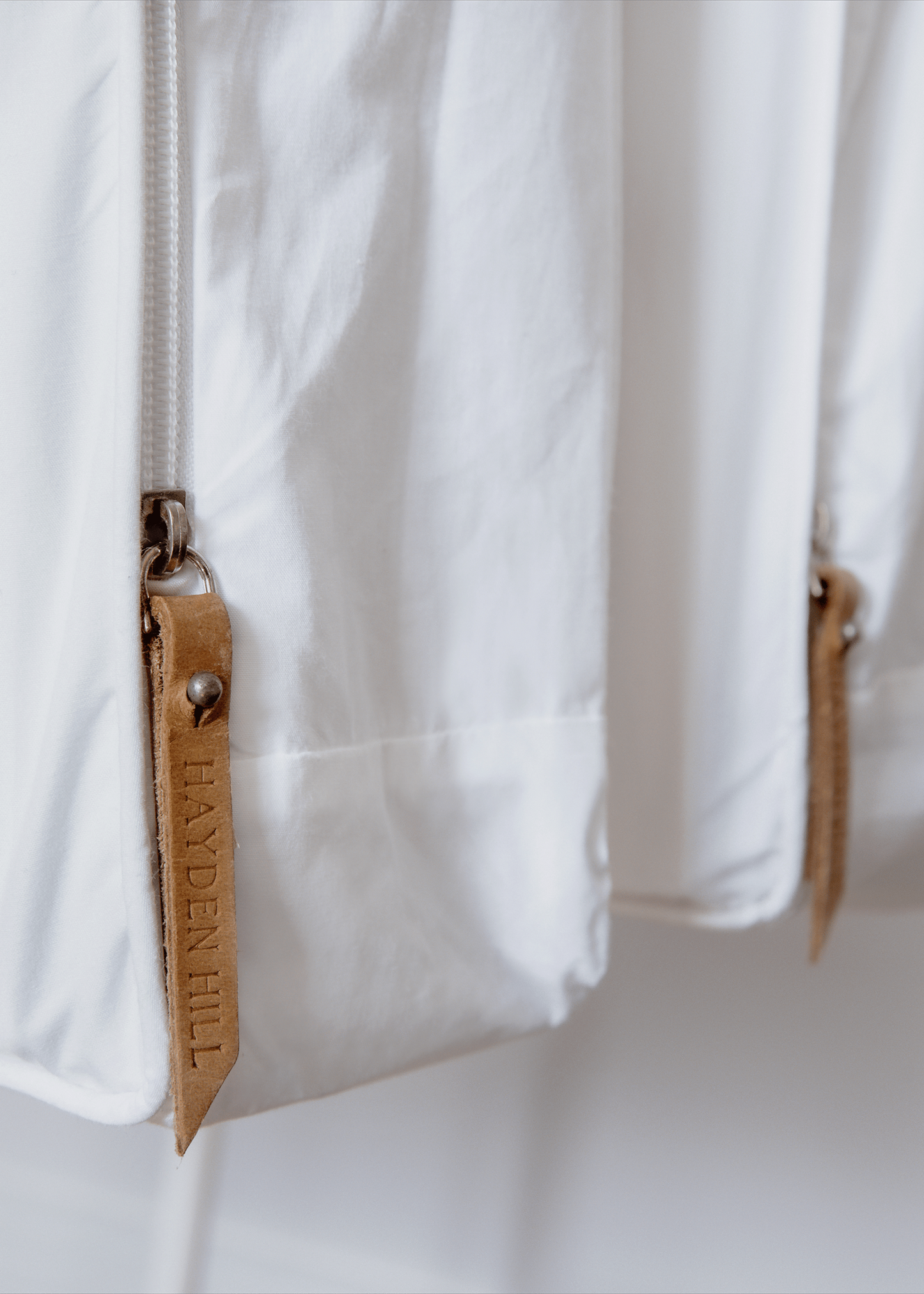 Close-up of a white garment bag with zipper and leather zipper pull branded with Hayden Hill logo. 