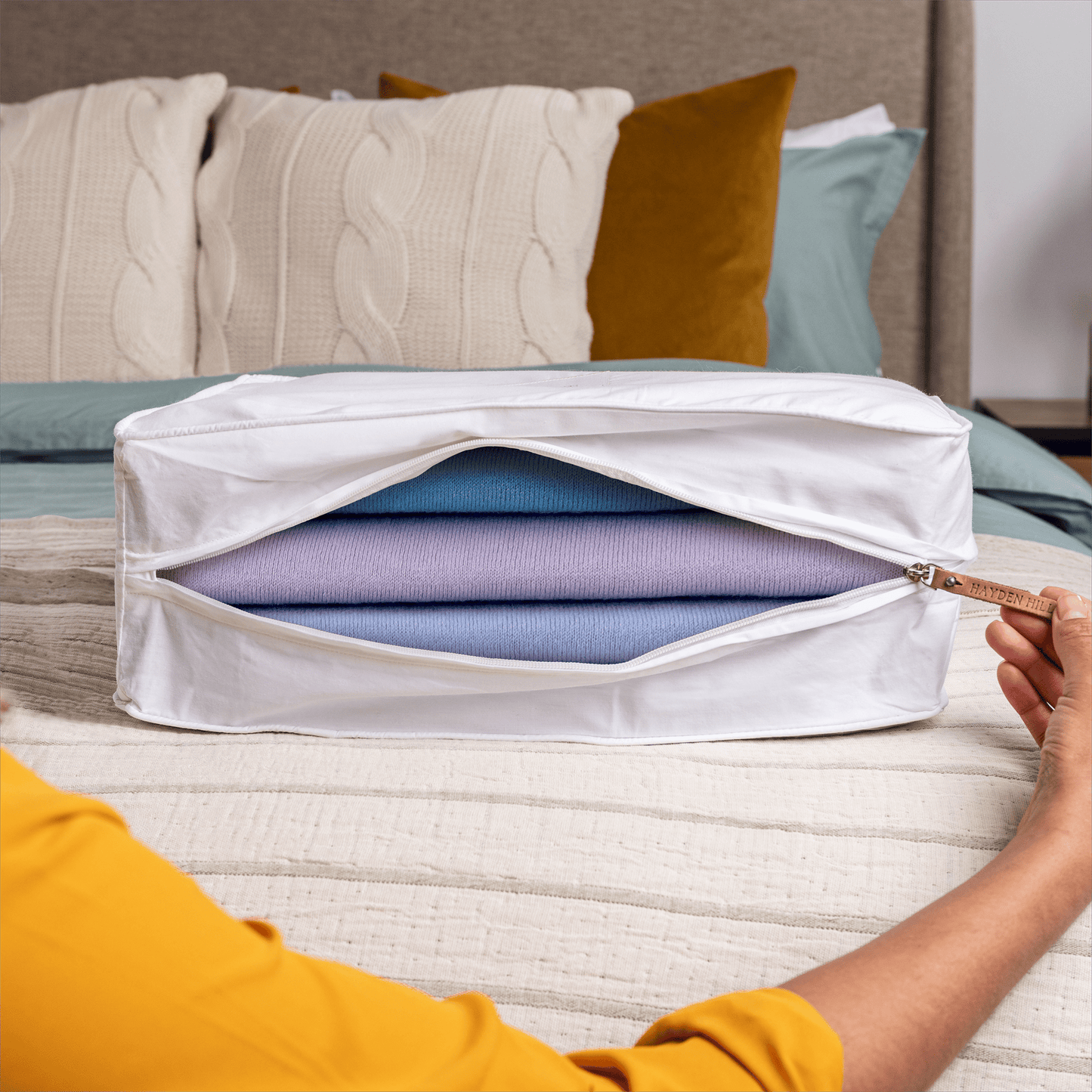 Person opening a white storage bag filled with folded clothes on a bed.