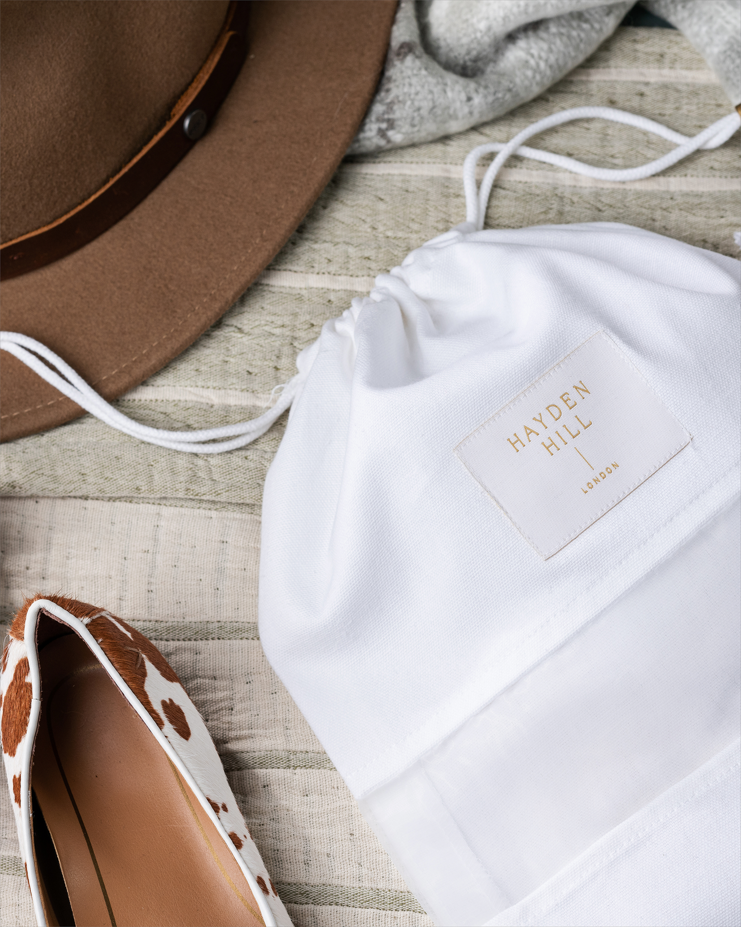 White drawstring bag with 'Hayden Hill' branding on a wooden surface with a brown hat and patterned shoes.