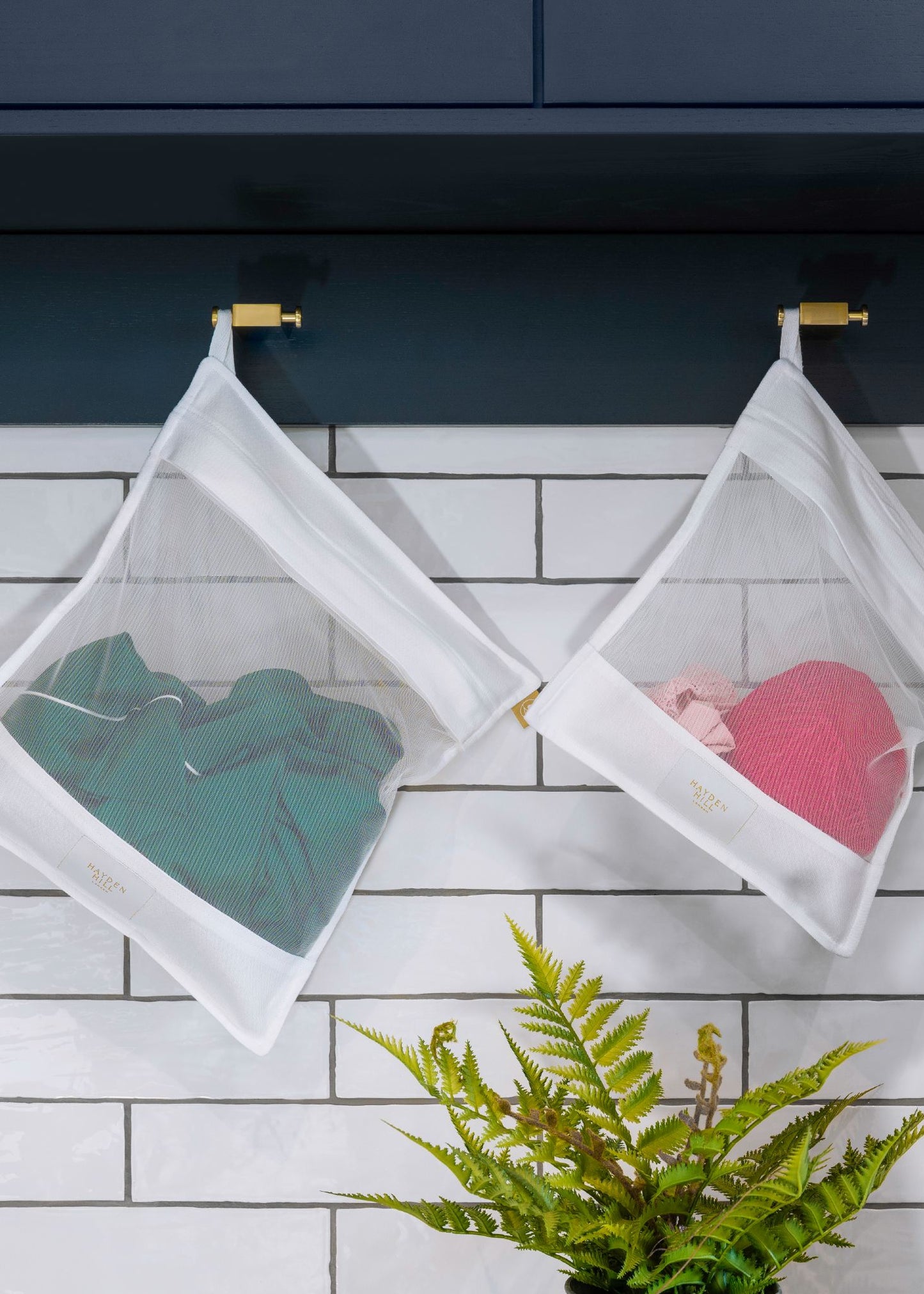 Clear mesh laundry bags hanging on a hook with clothes inside, on a tiled wall background.
