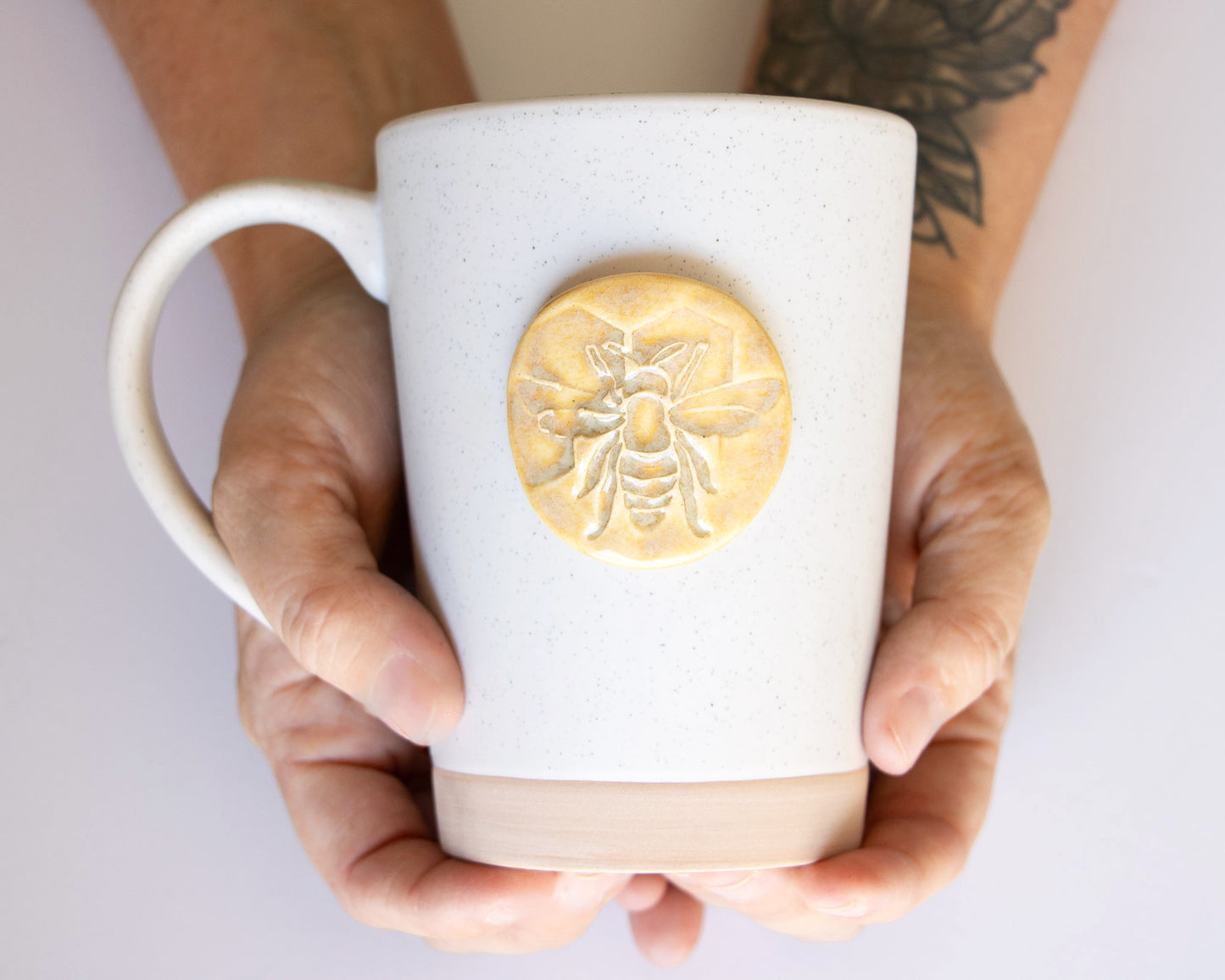 White mug with a gold bee emblem held by hands on a white background